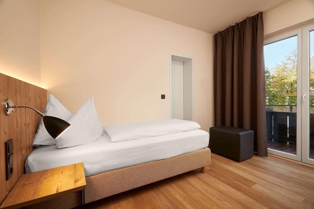 A modern hotel room with a bed, bedside table, and a view of the outdoors.