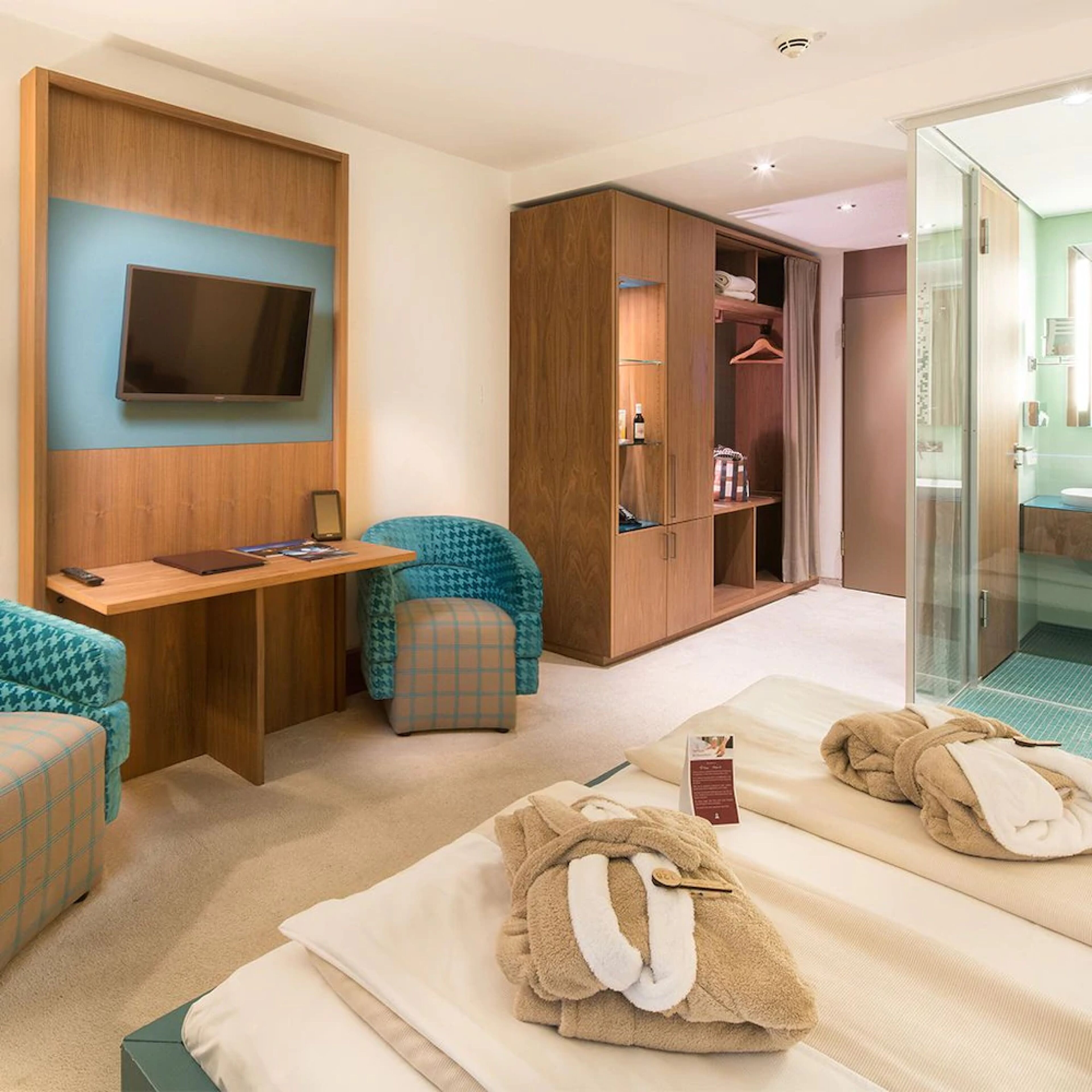 A hotel room with a TV mounted on the wall, a desk with a chair, a wardrobe, a glass shower area, and two beds with towels.
