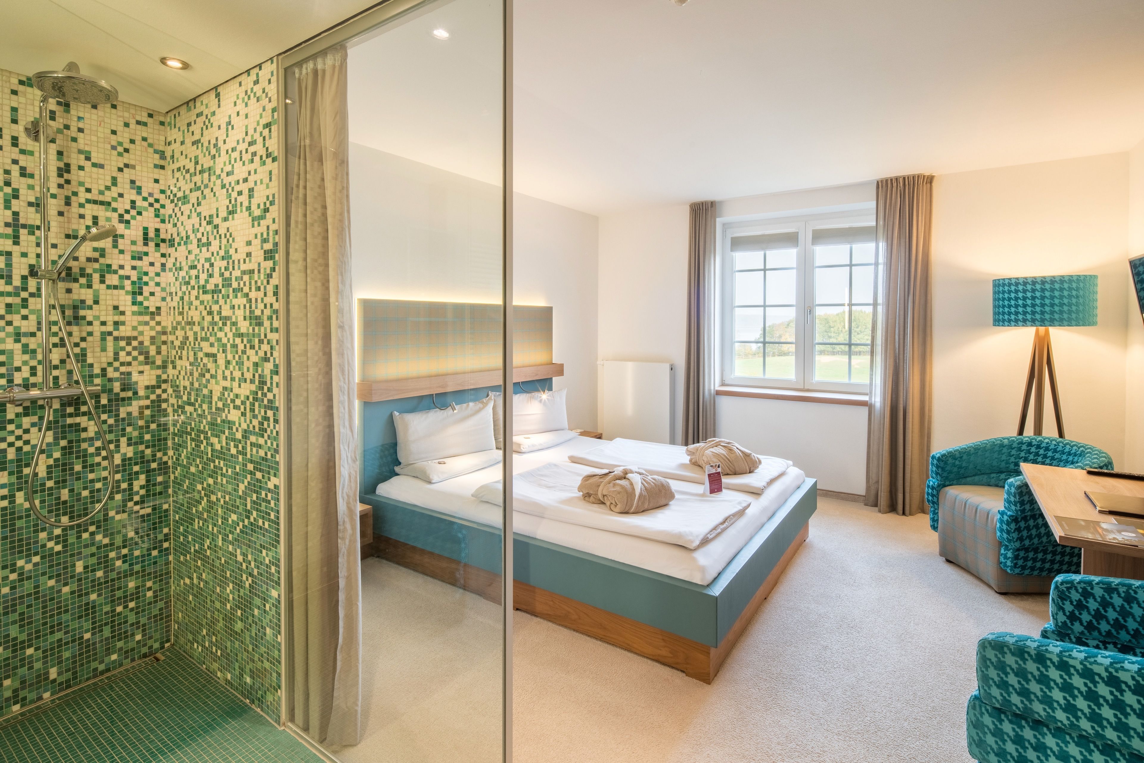 Ein Hotelzimmer mit einem großen Bett, einer Dusche mit Glastrennwand, einem Schreibtisch mit Lampe und Blick ins Freie.