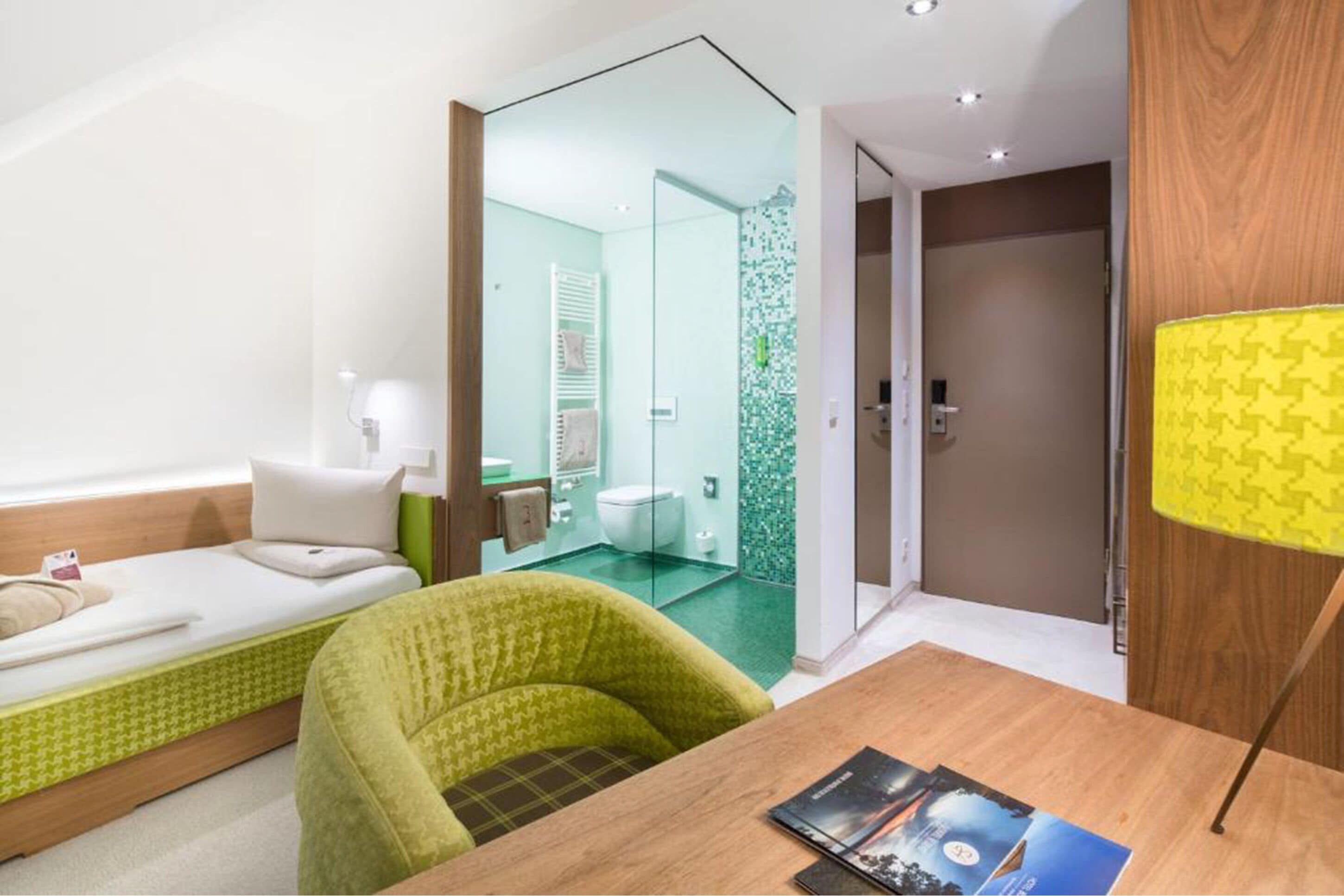 Ein modernes Hotelzimmer mit einem Bett, einem grünen Sessel, einem Holztisch mit Zeitschriften und einem durch eine Glastrennwand einsehbaren Badezimmer.