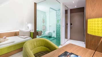 Ein modernes Hotelzimmer mit einem Bett, einem grünen Sessel, einem Holztisch mit Zeitschriften und einem durch eine Glastrennwand einsehbaren Badezimmer.