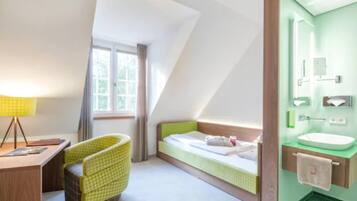 A hotel room with a single bed, a desk with a lamp, a chair, a window with curtains, and a bathroom with a sink and mirror.