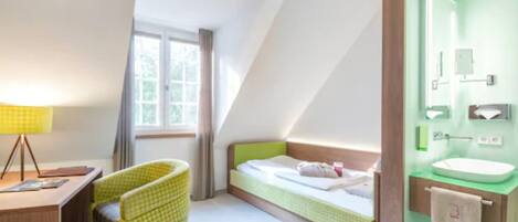 Une chambre d’hôtel avec un lit simple, un bureau avec une lampe, une chaise, une fenêtre avec des rideaux et une salle de bain avec un lavabo et un miroir.