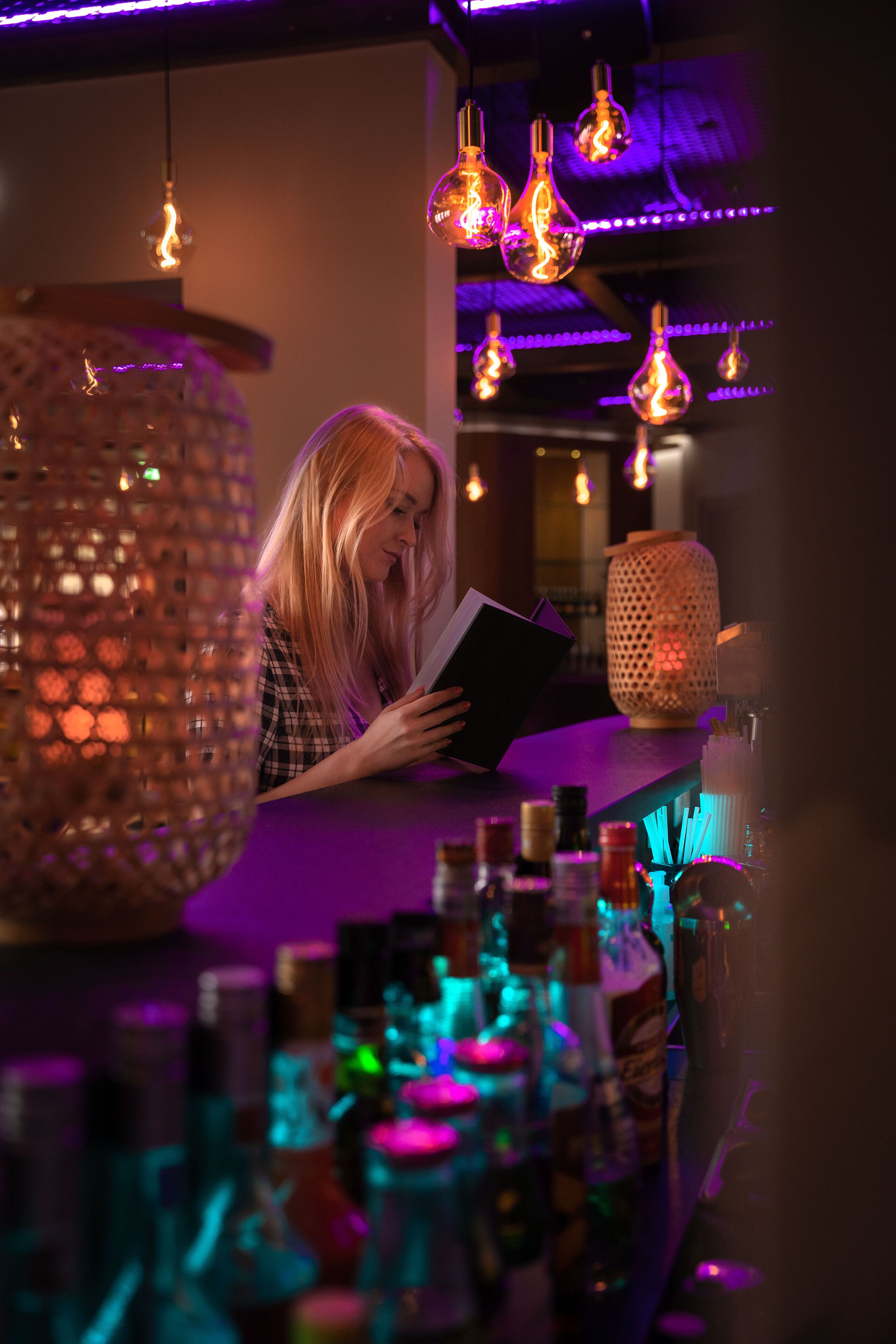 A person reading a book in a dimly lit bar with hanging light fixtures and a patterned wall.