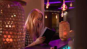 Una persona leyendo un libro en un bar tenuemente iluminado, con lámparas colgantes y un papel tapiz decorado.