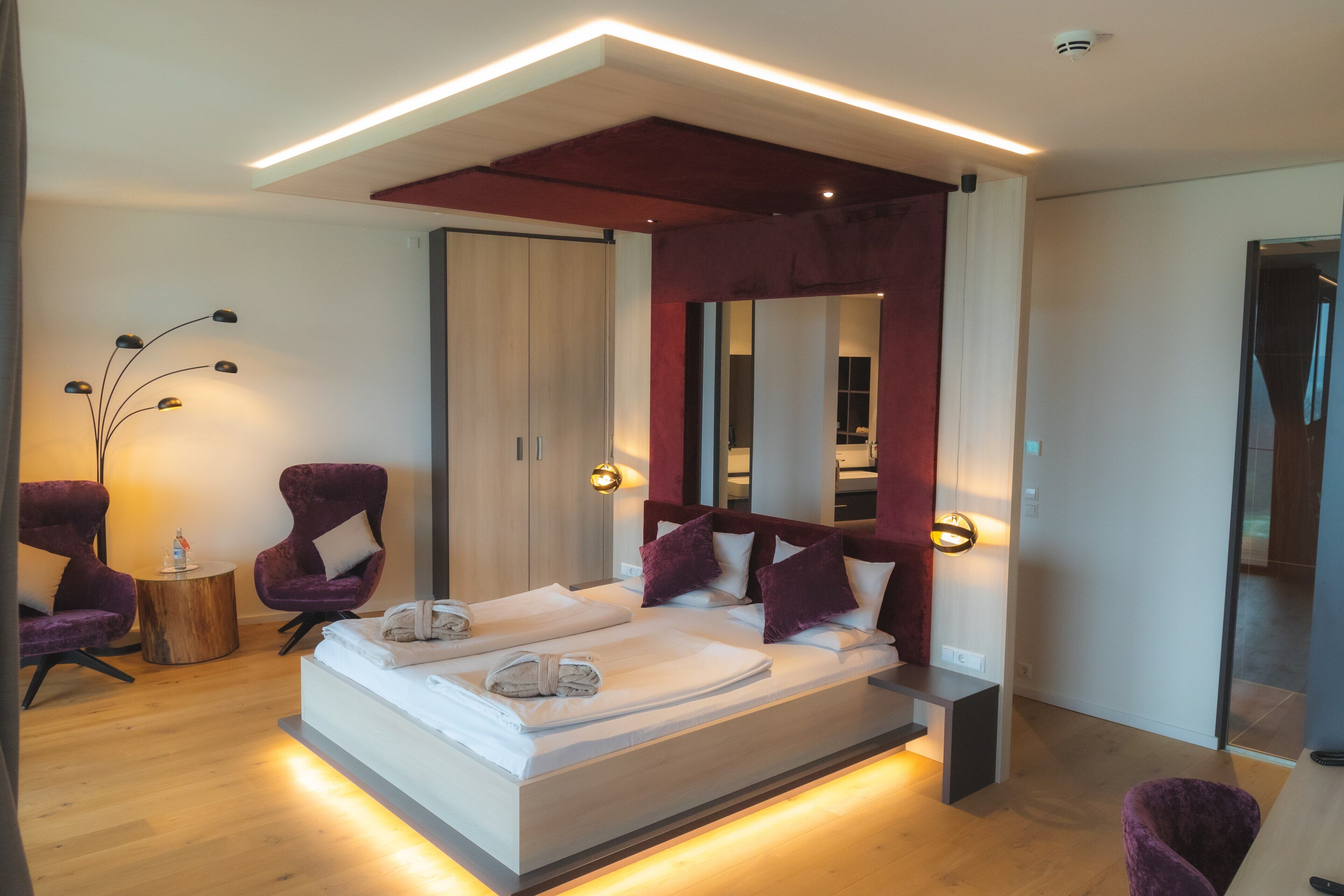A modern hotel room with a large bed, two purple armchairs, a round wooden side table, and a mirror above the bed.
