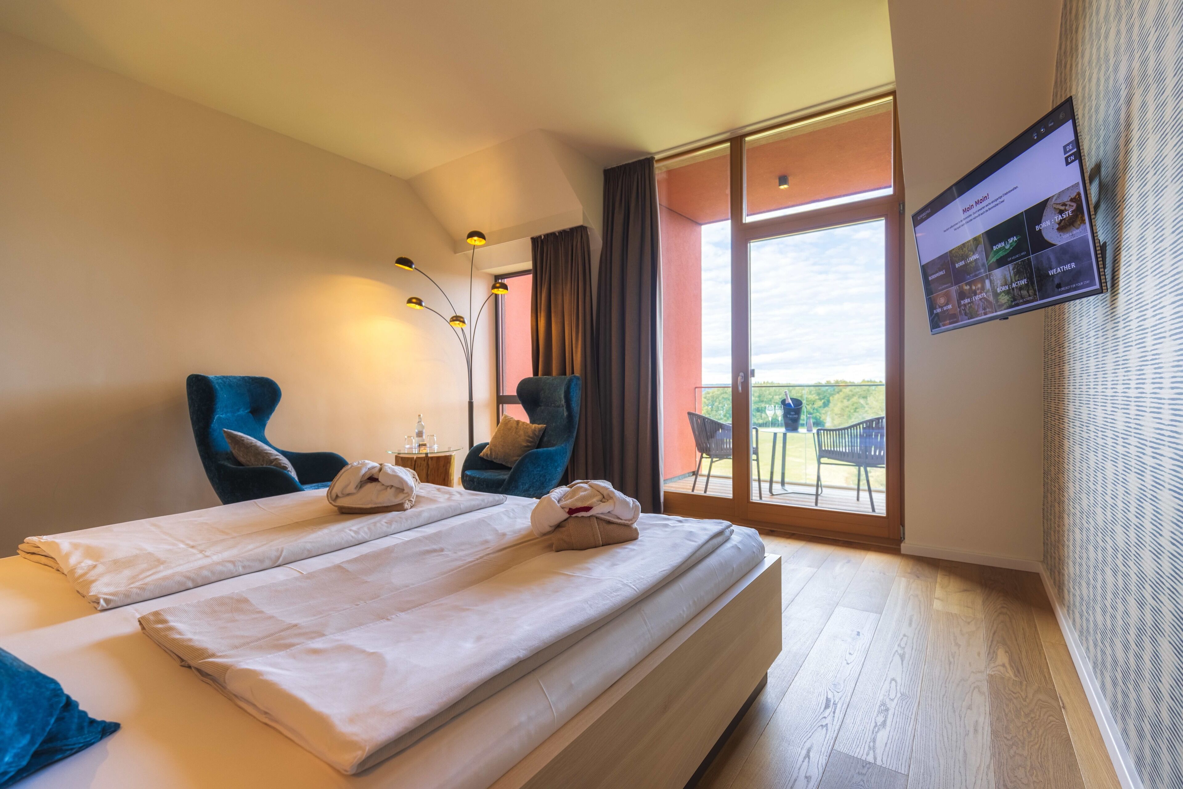 A hotel room with a large bed, two chairs, a table with a lamp, and a view of the outdoors through a large window.