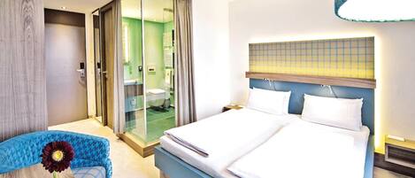 A hotel room with a large bed, a seating area with a chair, and a glass door leading to a bathroom.