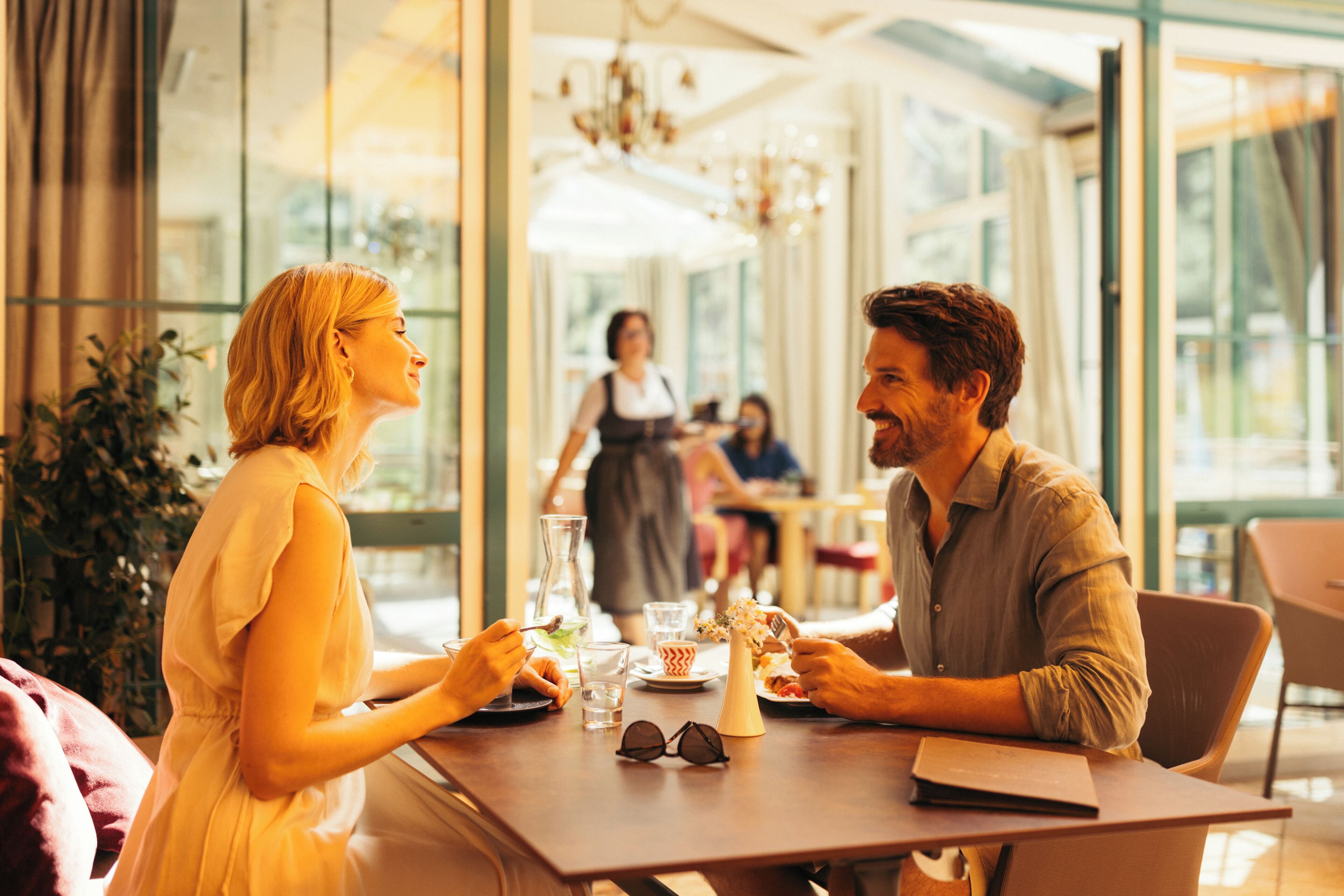 A couple dining at a table with a view of the outdoors, a waiter in the background, and a casual dining atmosphere.
