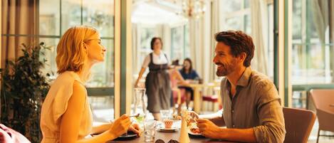 A couple dining at a table with a view of the outdoors, a waiter in the background, and a casual dining atmosphere.