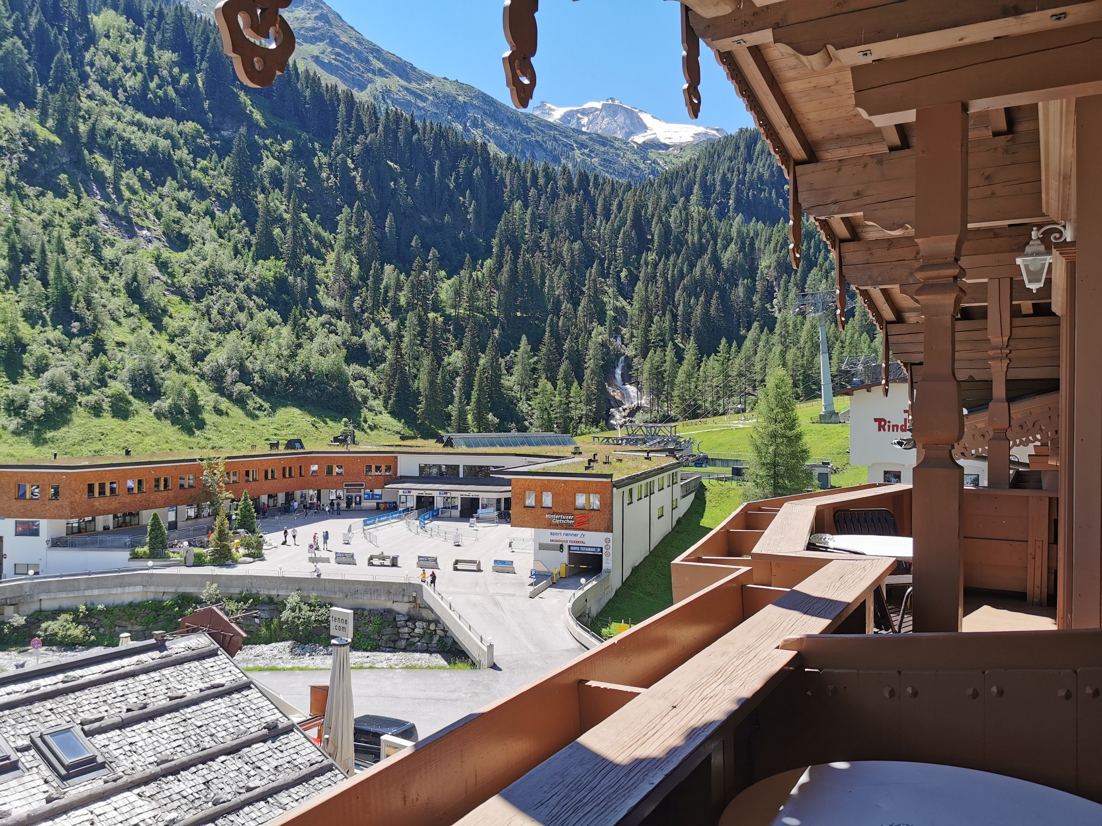 Vista da un balcone in legno su una zona montana con un edificio che include un centro sportivo e un parcheggio.