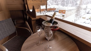 Et træbord med en champagneflaske og to glas, en stol og udsigt til et sneklædt landskab gennem et vindue.