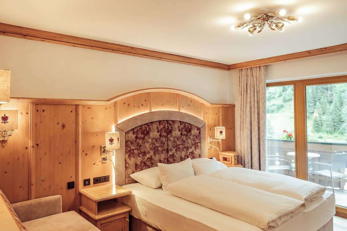 Un dormitorio con una cama grande, cabecera de madera, mesita de noche, sofá y un amplio ventanal con vistas a zonas verdes.