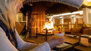 En person holder en drink i hyggelige omgivelser med en synlig pejs og møbler af træ.