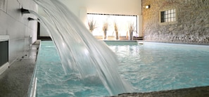 An indoor swimming pool with a waterfall feature, stone walls, and potted plants.