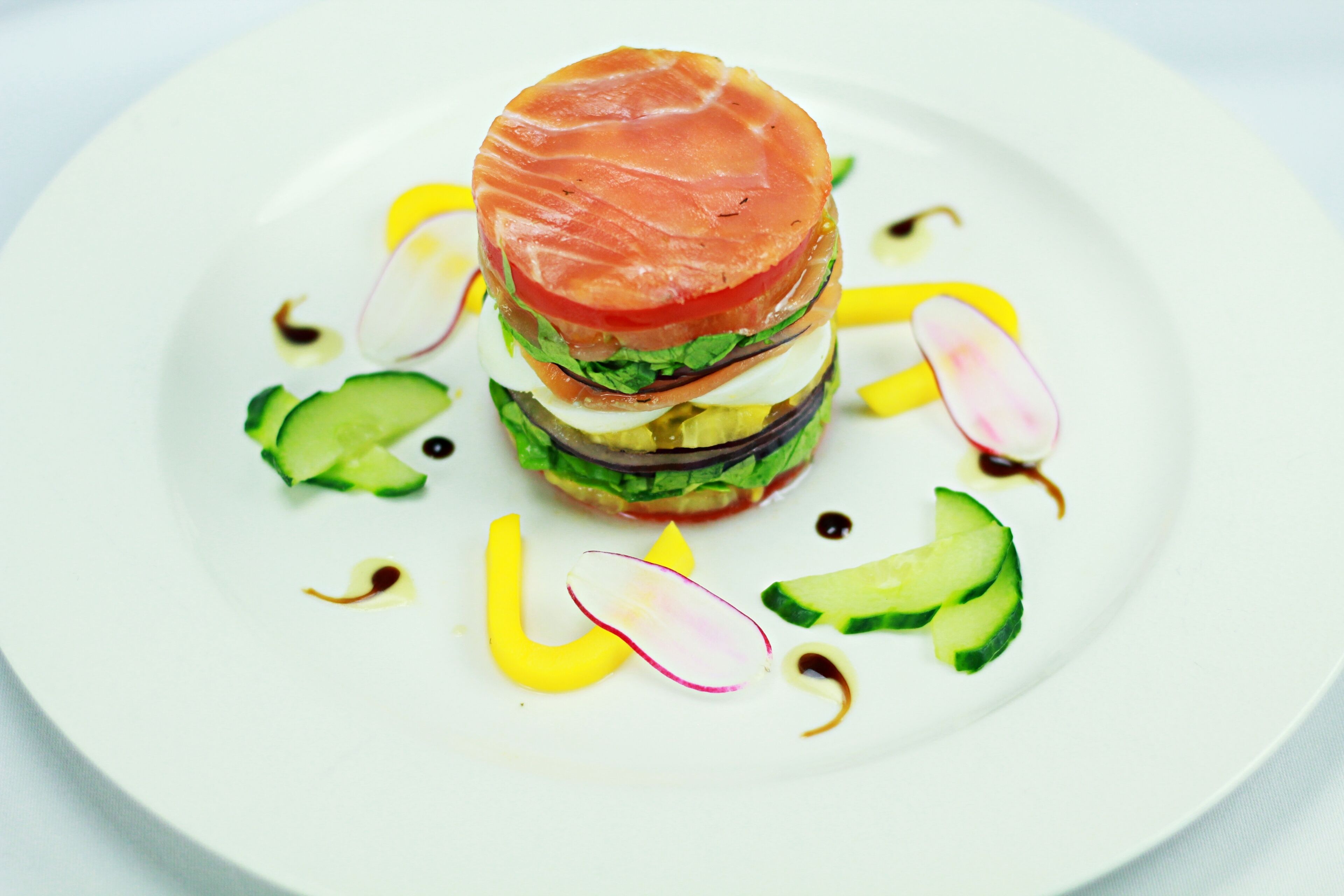 A white plate with a layered sandwich, cucumber slices, radish slices, and yellow sauce.