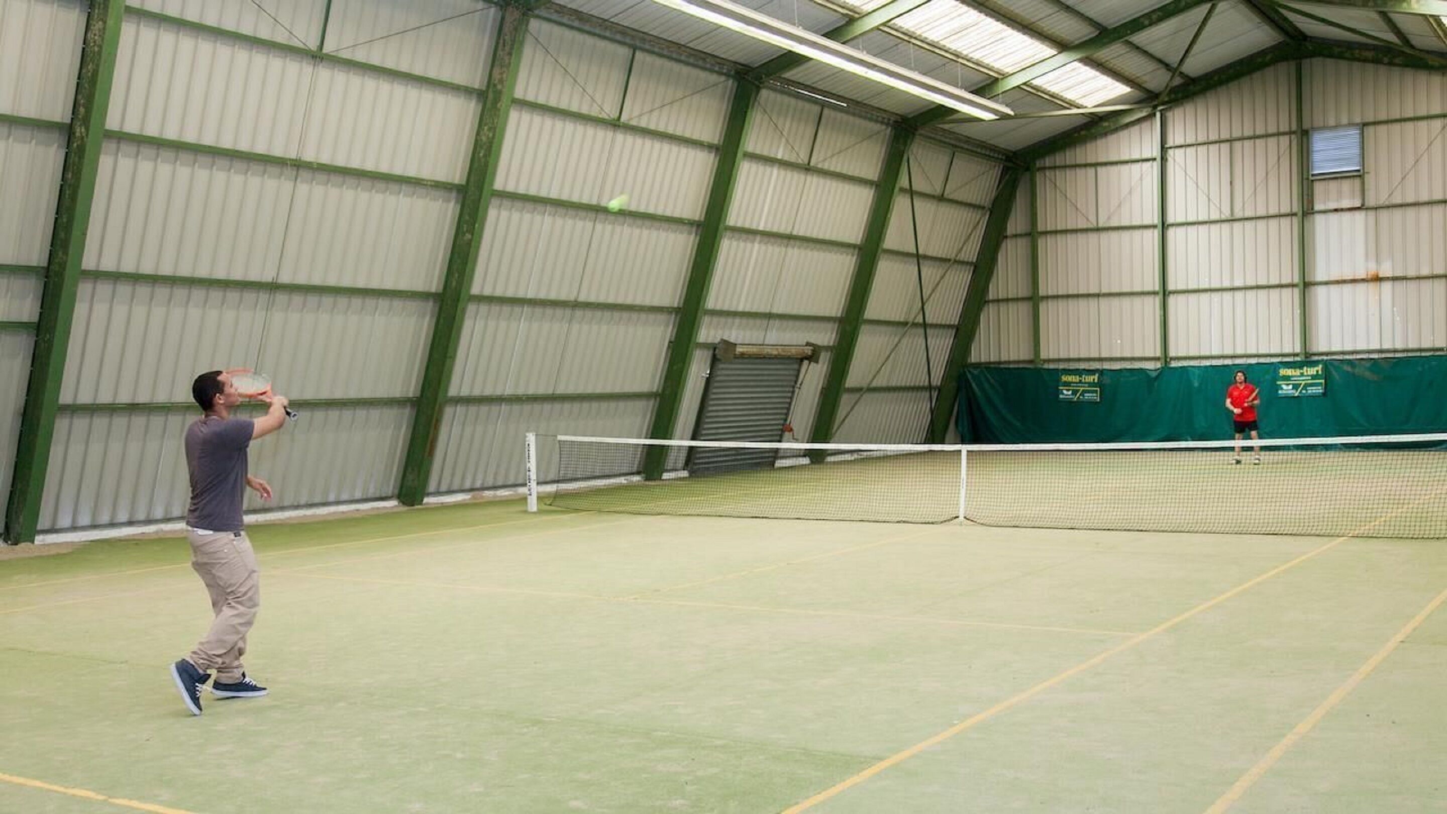 An indoor tennis court with a net, a player in the foreground, and another player in the background.