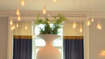 A dining area with tables set for service, a central white vase with greenery, and hanging light bulbs.