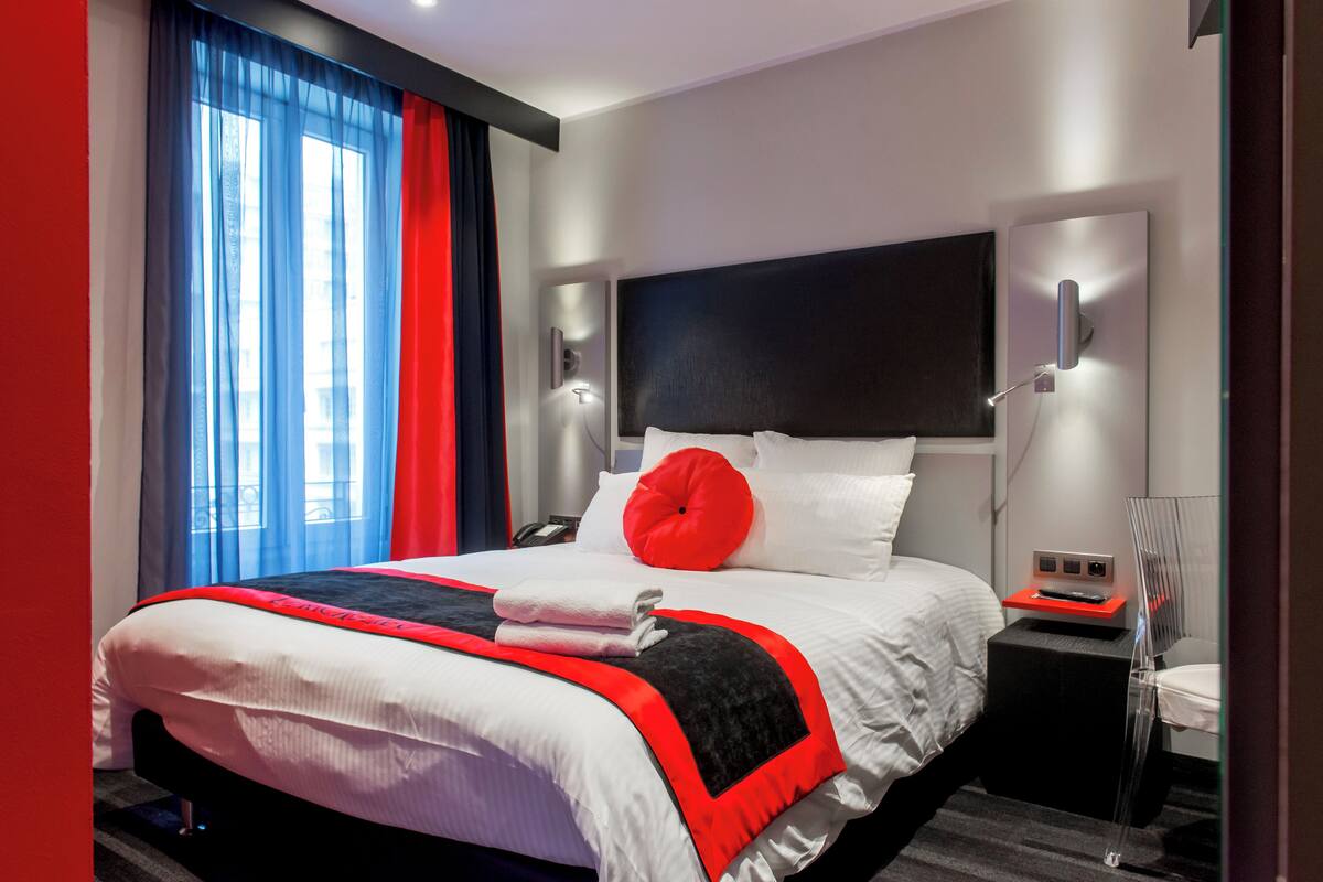 Une chambre d’hôtel moderne avec un grand lit, une table de chevet avec une lampe et une vue sur la ville grâce à de grandes fenêtres.