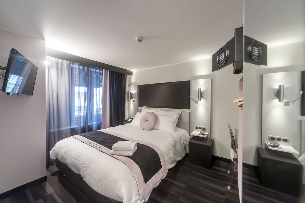 Une chambre d’hôtel moderne dotée d’un grand lit, de tables de chevet, d’un téléviseur à écran plat et d’une grande fenêtre avec des rideaux.