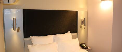Una habitación de hotel con cama, mesita de noche, teléfono y lámpara fijada a la pared.