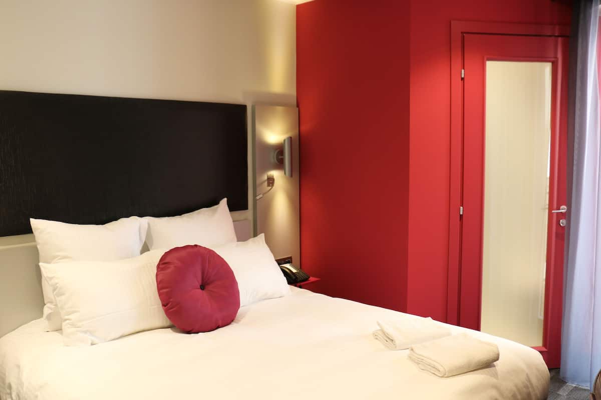 Une chambre d’hôtel avec un lit, du linge de lit blanc, un mur rouge et une porte.