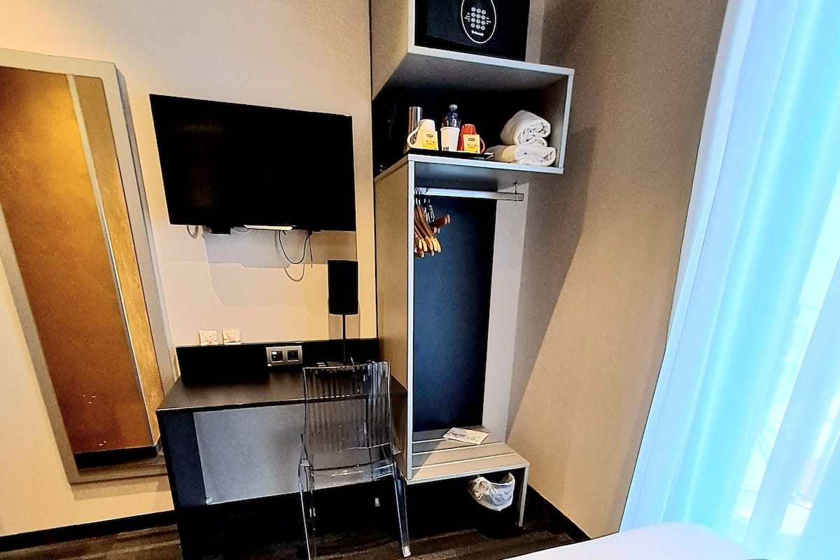 Une chambre d’hôtel avec un lit, un téléviseur fixé au mur, un petit bureau avec une chaise, une étagère avec des articles de toilette et une fenêtre avec des rideaux.