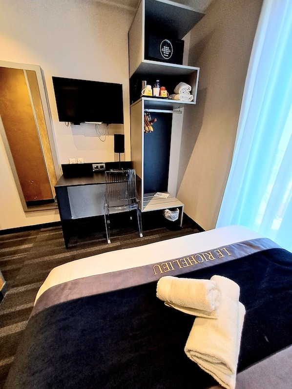 A hotel room with a bed, a TV mounted on the wall, a small desk with a chair, a shelf with toiletries, and a window with curtains.