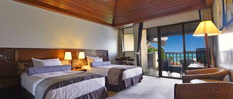 Habitación de hotel con dos camas, un ventilador de techo, un balcón con mobiliario de mimbre y vista al océano.