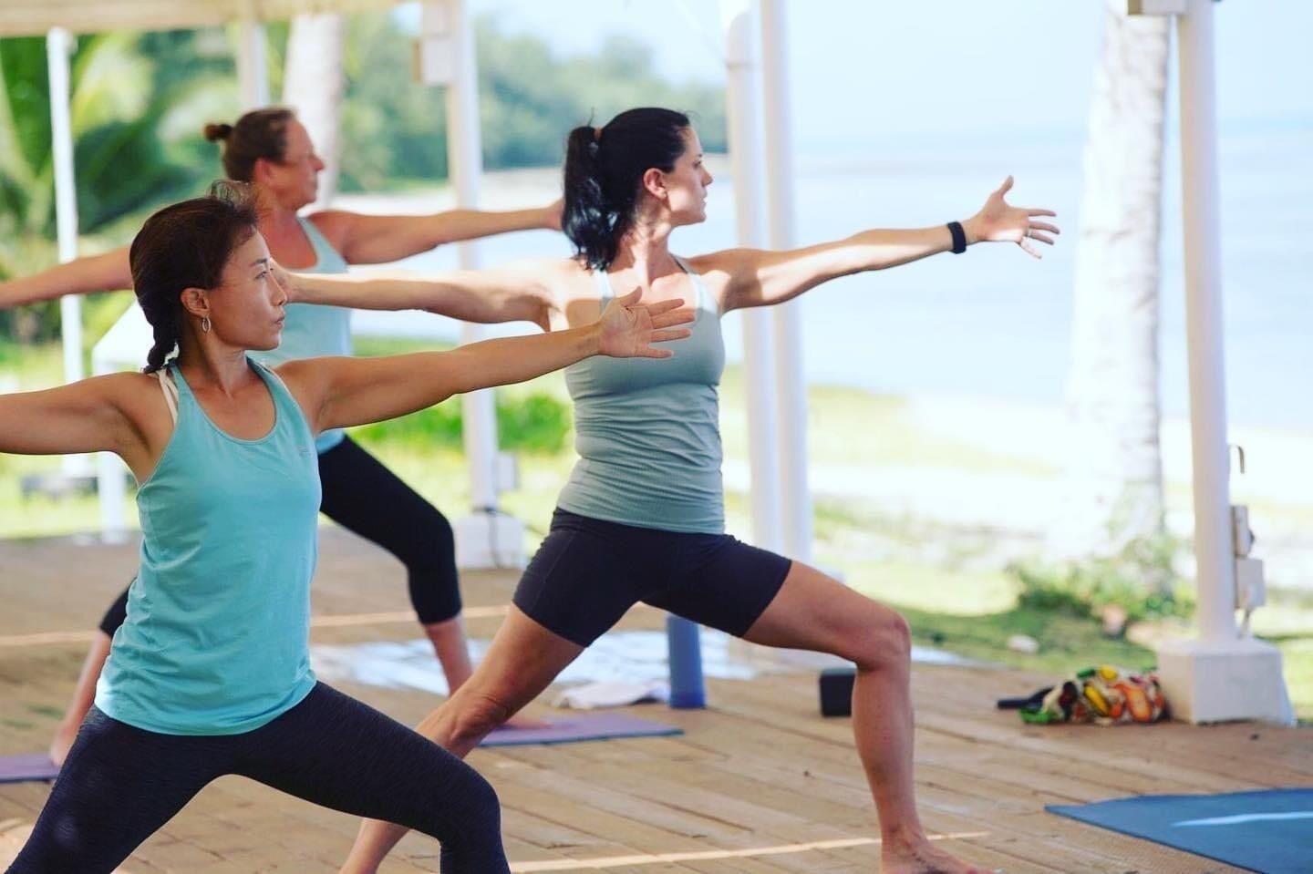Un grupo de personas en una clase de yoga al aire libre, con piso de madera y colchonetas de ejercicio.
