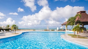 A large, clear swimming pool with lounge chairs and umbrellas, a palm tree, and a view of the ocean. - Aqua Resort Club Saipan (Saipan)