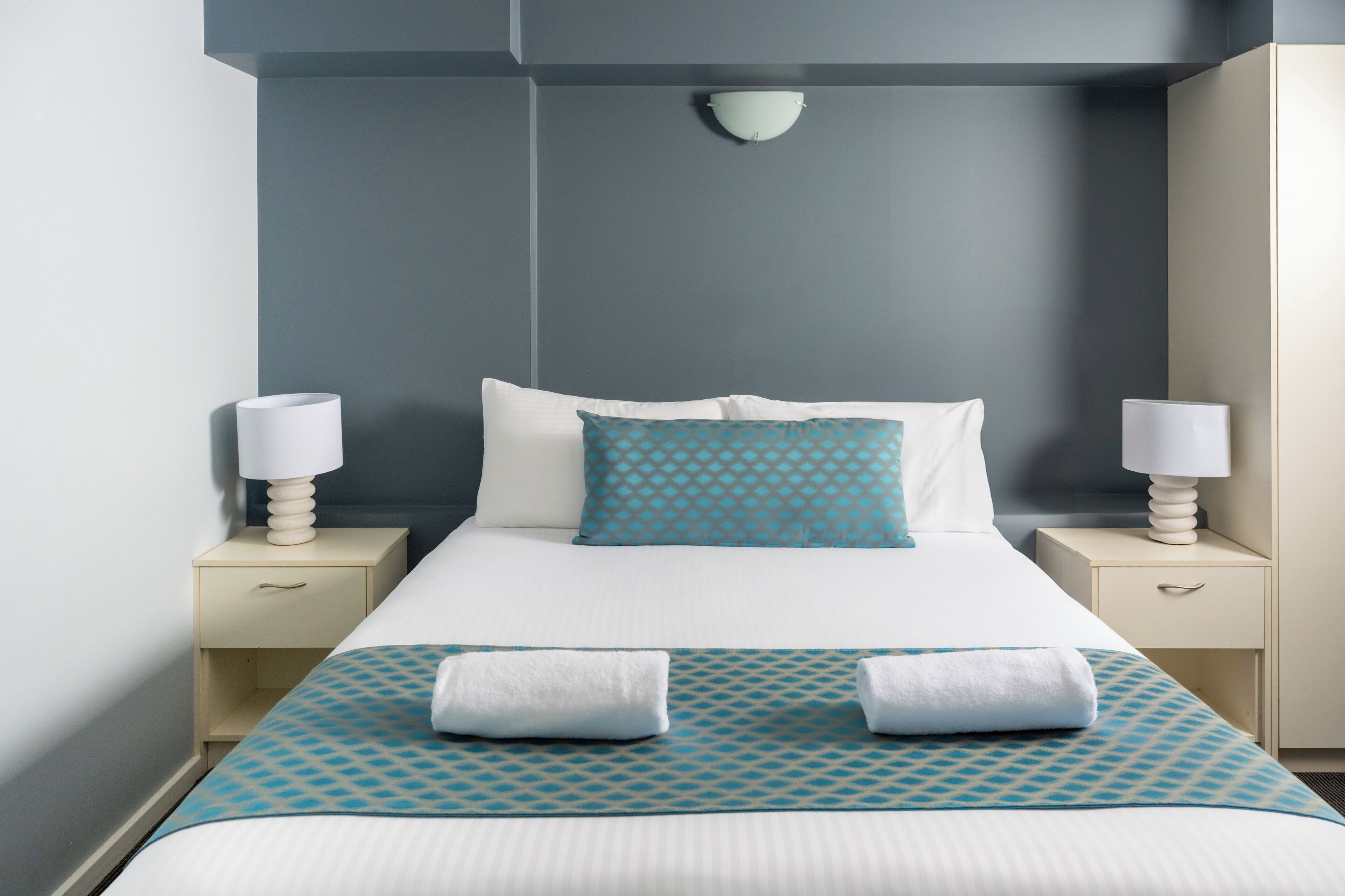 Una cama bien hecha con ropa de cama blanca y azul, dos lámparas de noche y un aplique de pared.