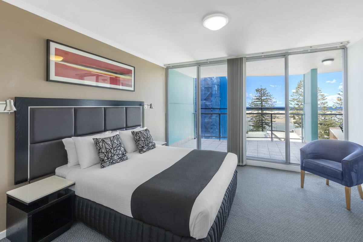 Een moderne hotelkamer met een groot bed, nachtkastje, stoel en een balkon met uitzicht op de omgeving.