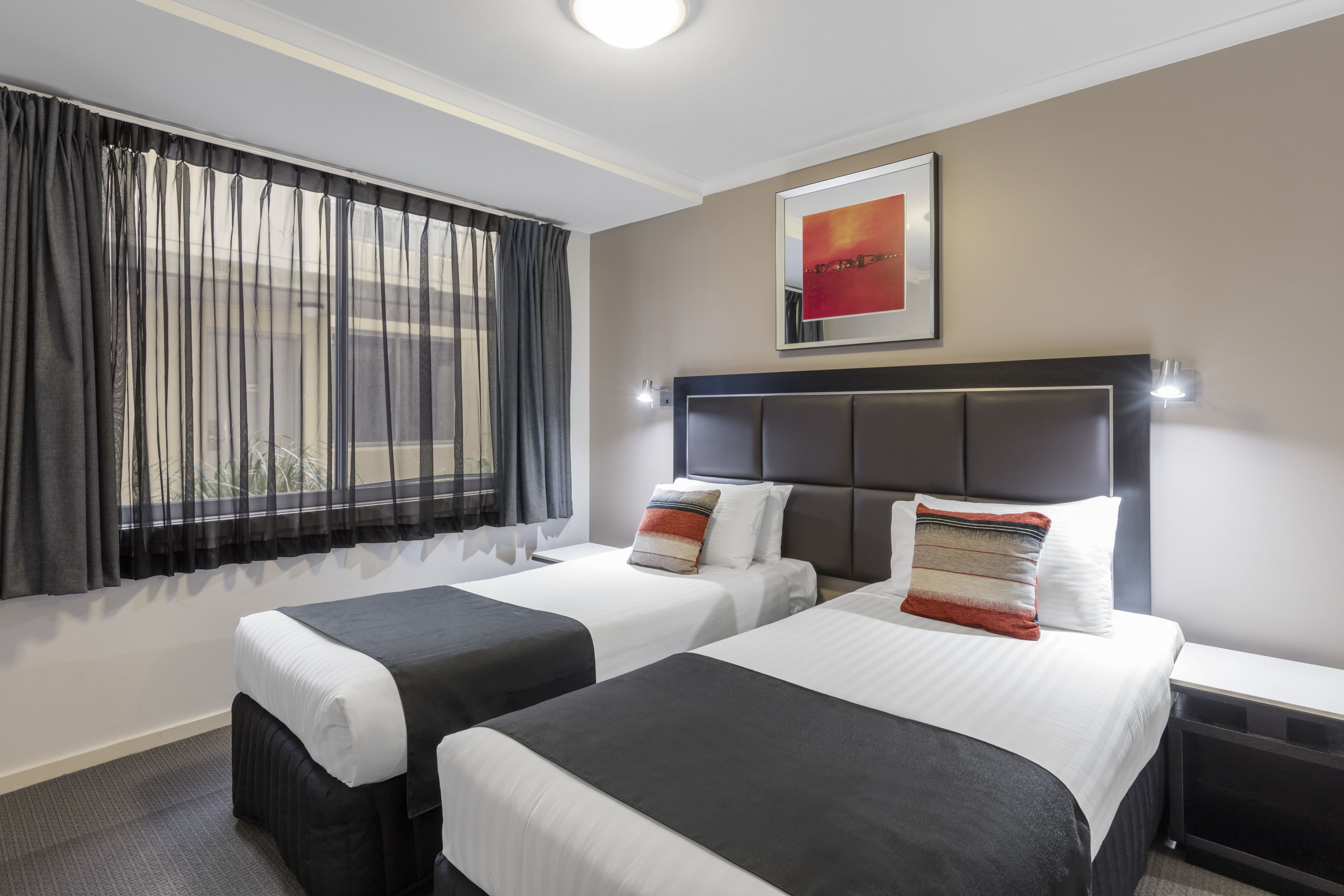Une chambre d’hôtel avec deux lits, une grande fenêtre avec des rideaux légers, une tête de lit dotée d’un luminaire et un tableau encadré au mur.
