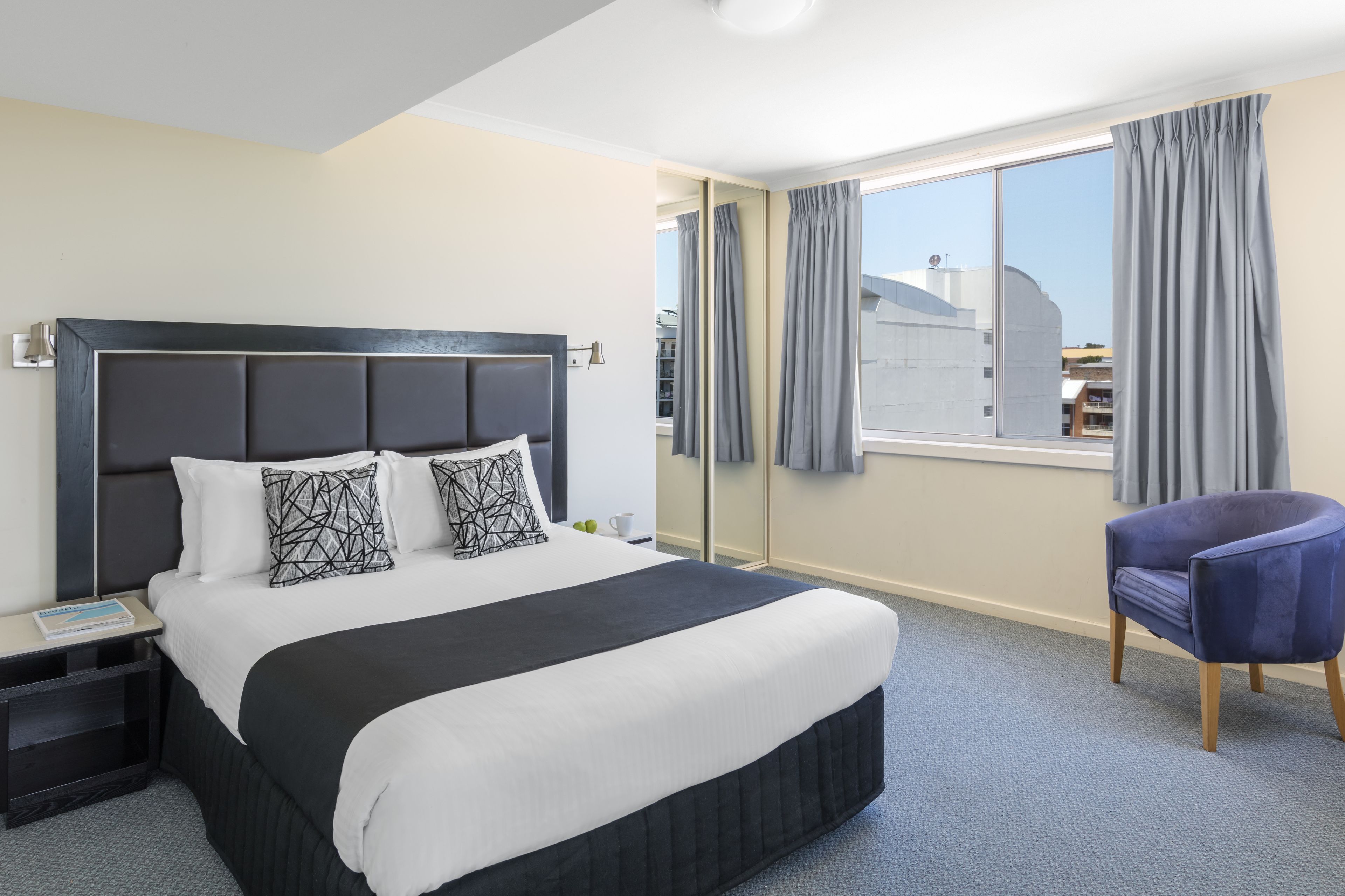 Una habitación de hotel con una cama grande, una mesita de noche, una silla y un ventanal con cortinas.