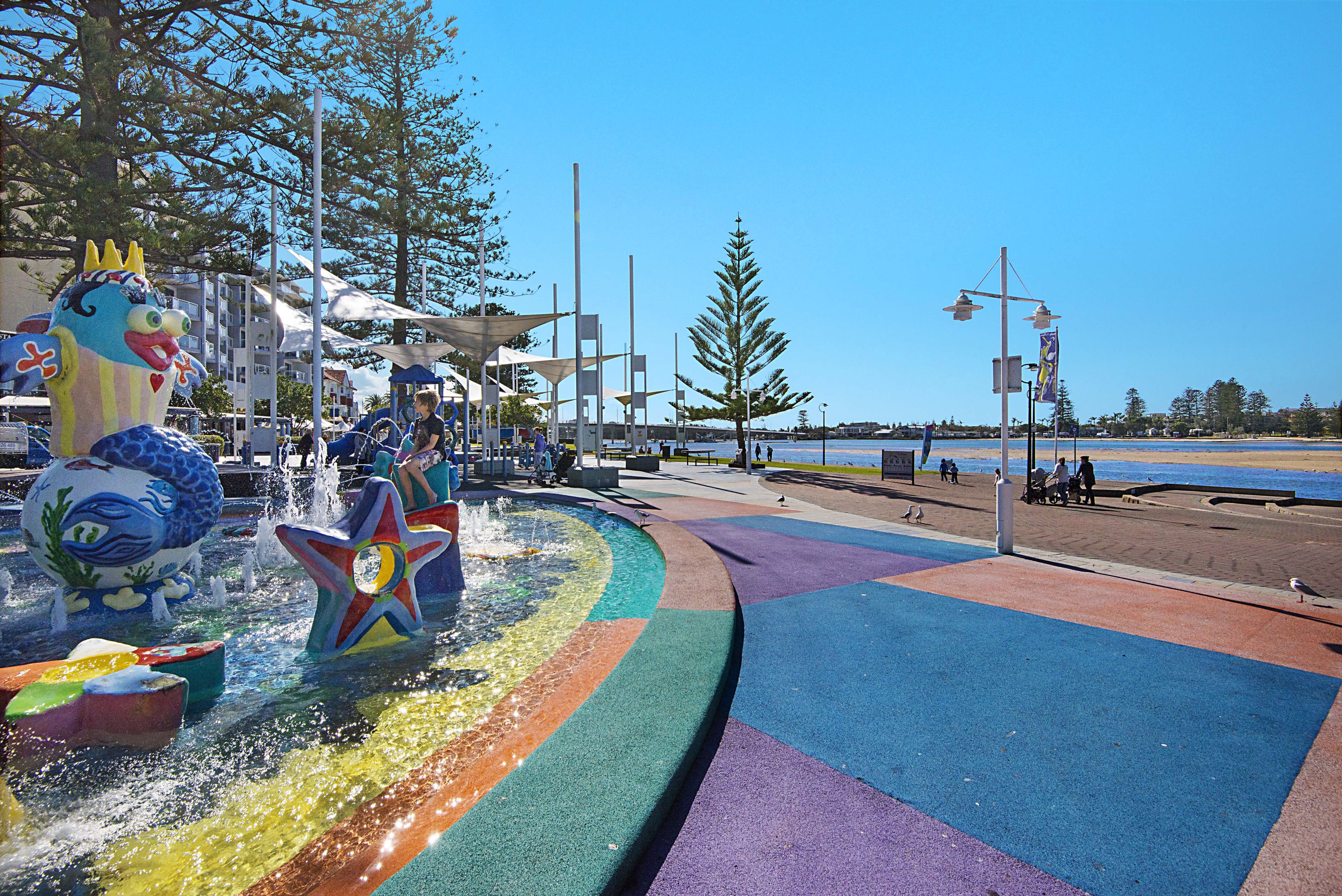 A colorful water fountain with a dragon and starfish design, surrounded by a playground area with various play structures.