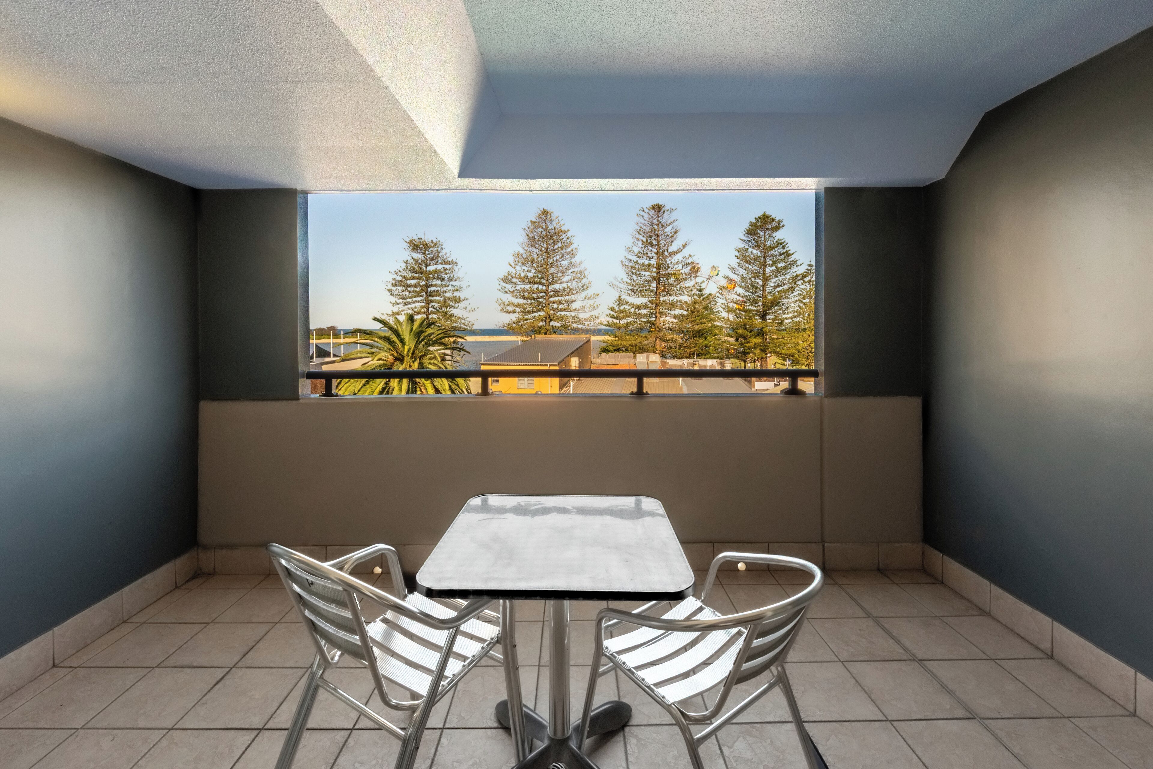 A balcony with a table and chairs, a view of trees and a building, and a tiled floor.