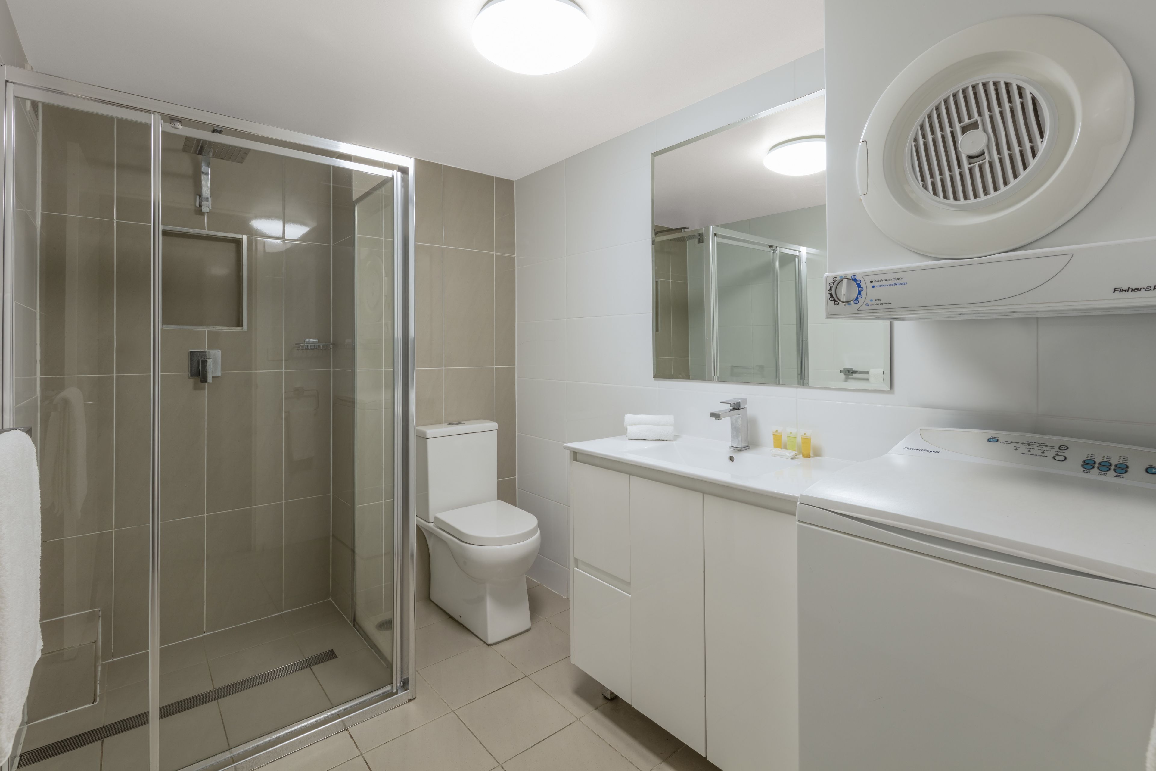 A modern bathroom with a glass shower enclosure, a toilet, and a white sink with a Fisher & Paykel washing machine.