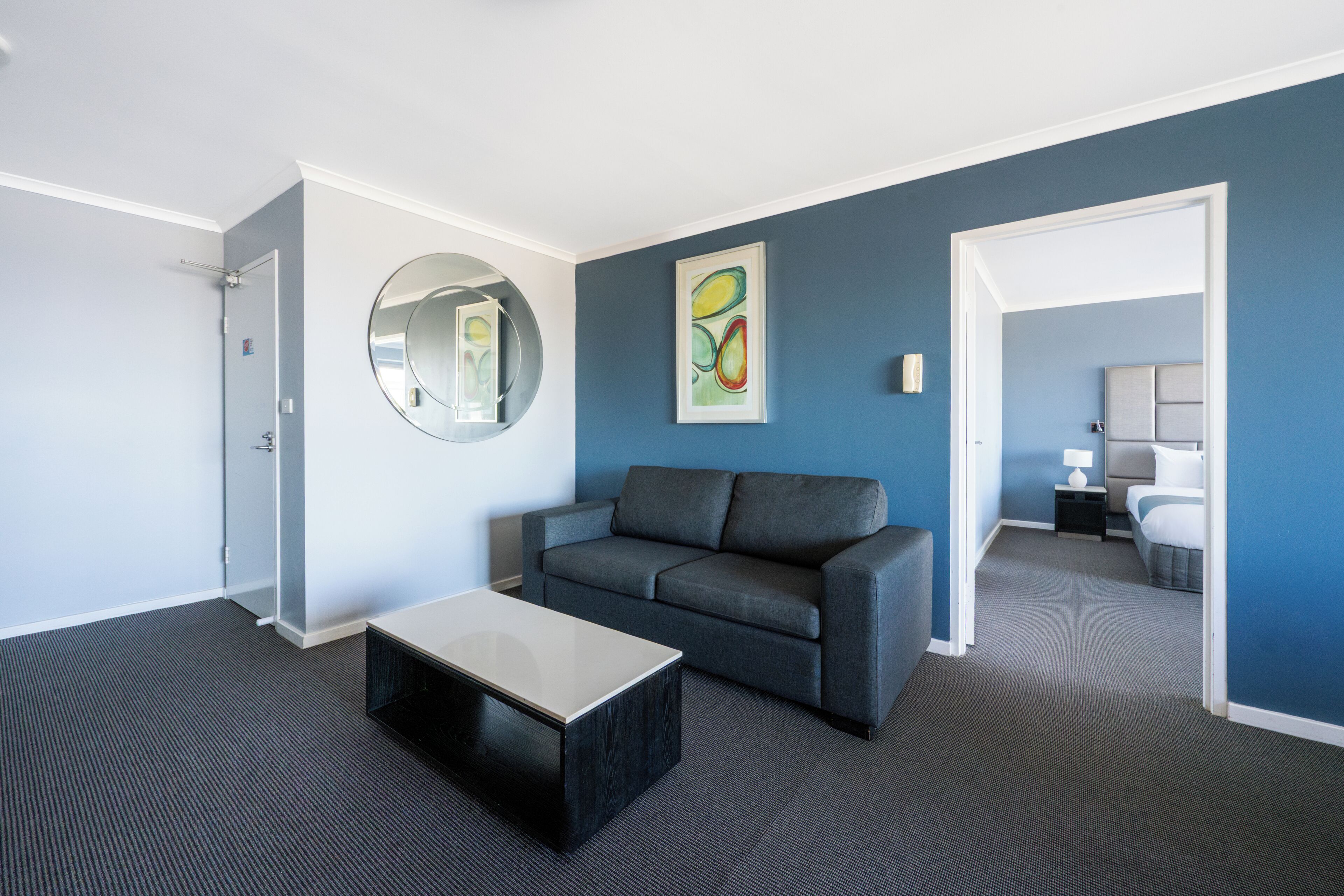 Une chambre d’hôtel moderne avec un canapé gris, un miroir rond et un tableau accroché au mur.