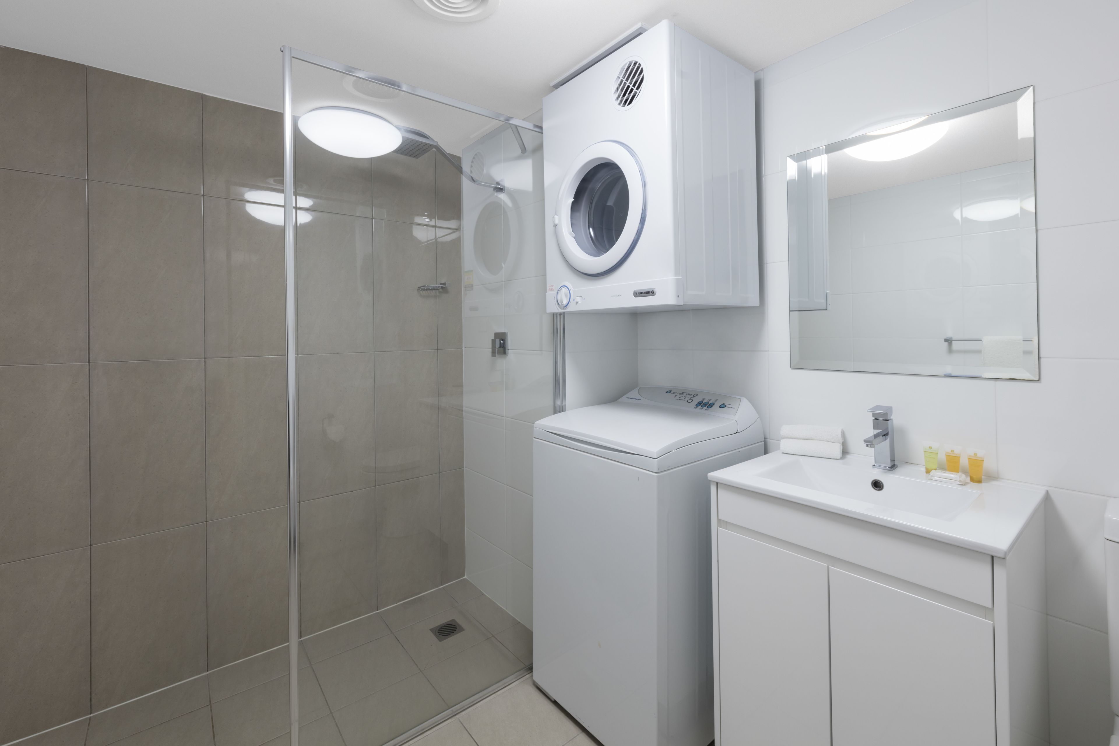 Un baño moderno con cabina de ducha de vidrio, lavadora integrada y lavamanos con espejo superior.