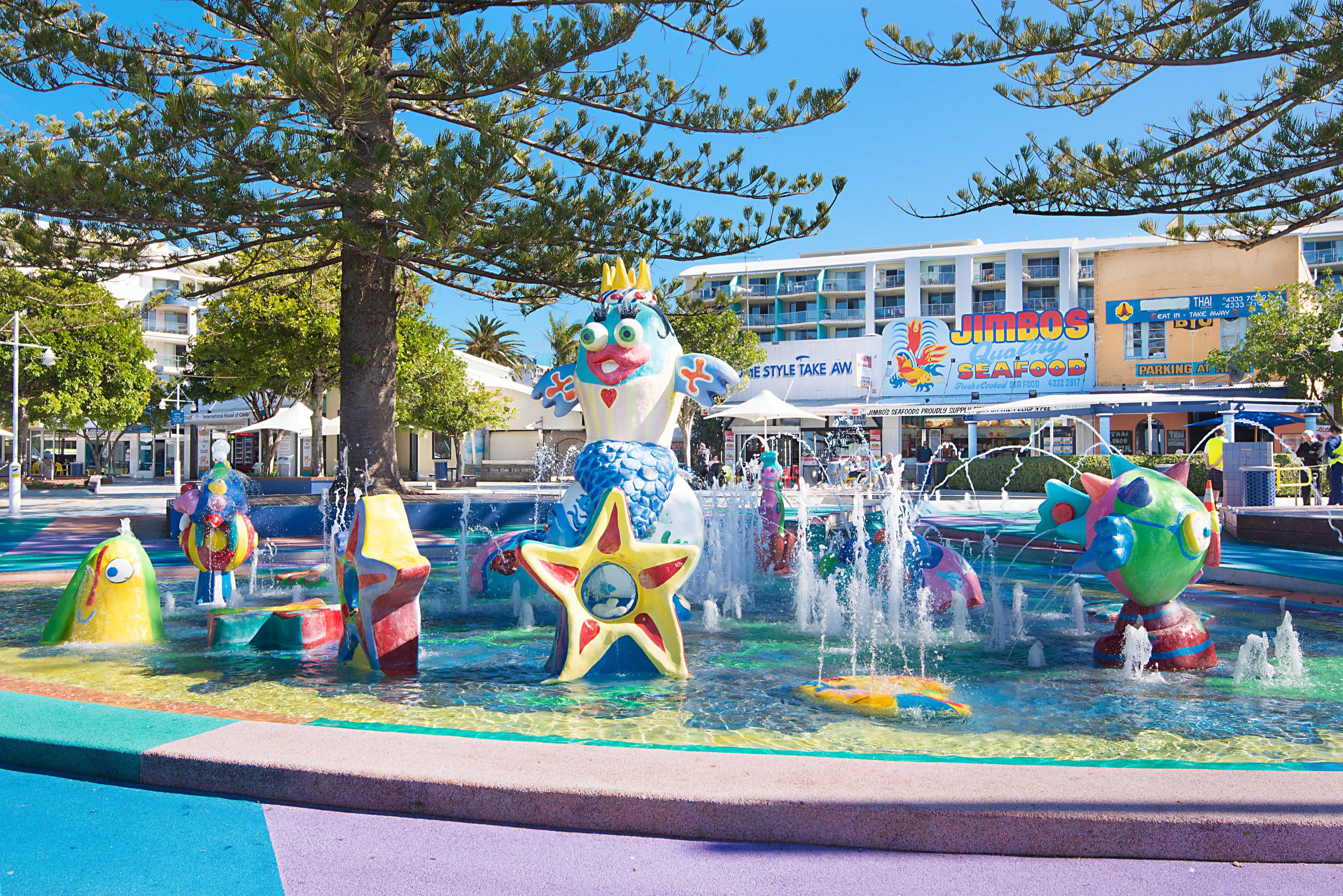 A colorful water fountain with various animal sculptures, a large starfish, and a building with a seafood restaurant in the background.