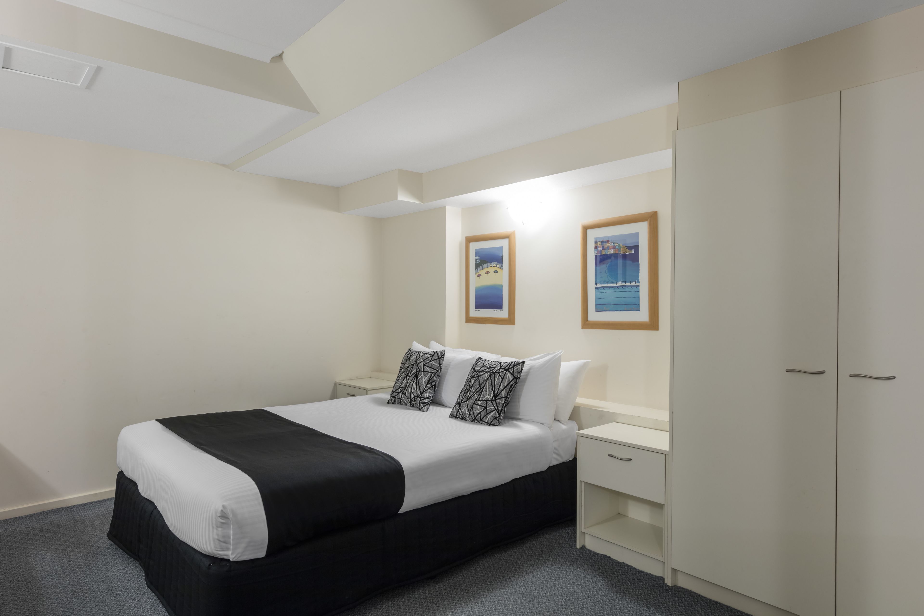 A hotel room with a bed, a wardrobe, a nightstand, and two framed pictures on the wall.