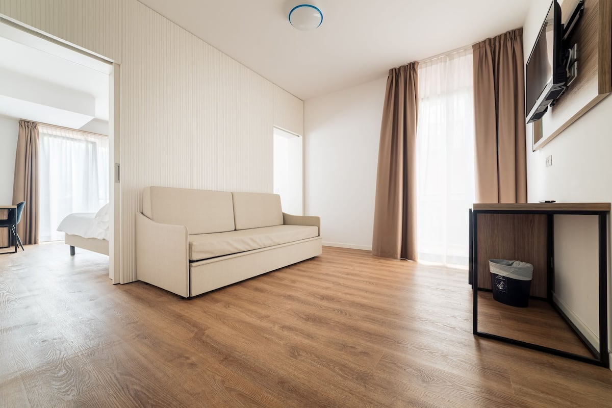 A modern living room with a beige sofa, wooden flooring, a TV on a wooden stand, and a large window with curtains.