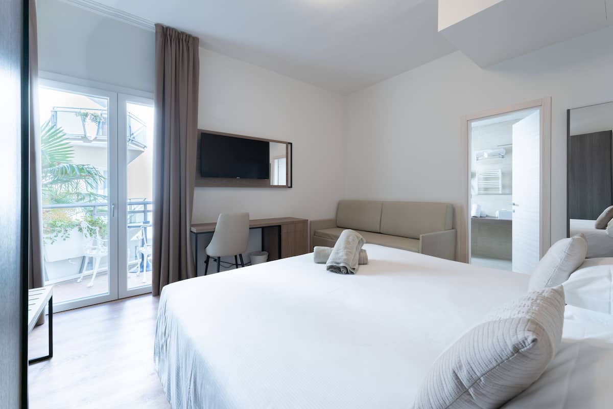 A modern hotel room with a large bed, a desk, a chair, a TV, and a balcony with plants.