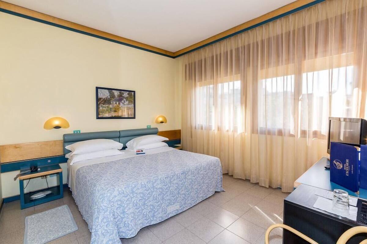 A hotel room with a bed, two bedside lamps, a nightstand, a desk with a computer, and a window with curtains.