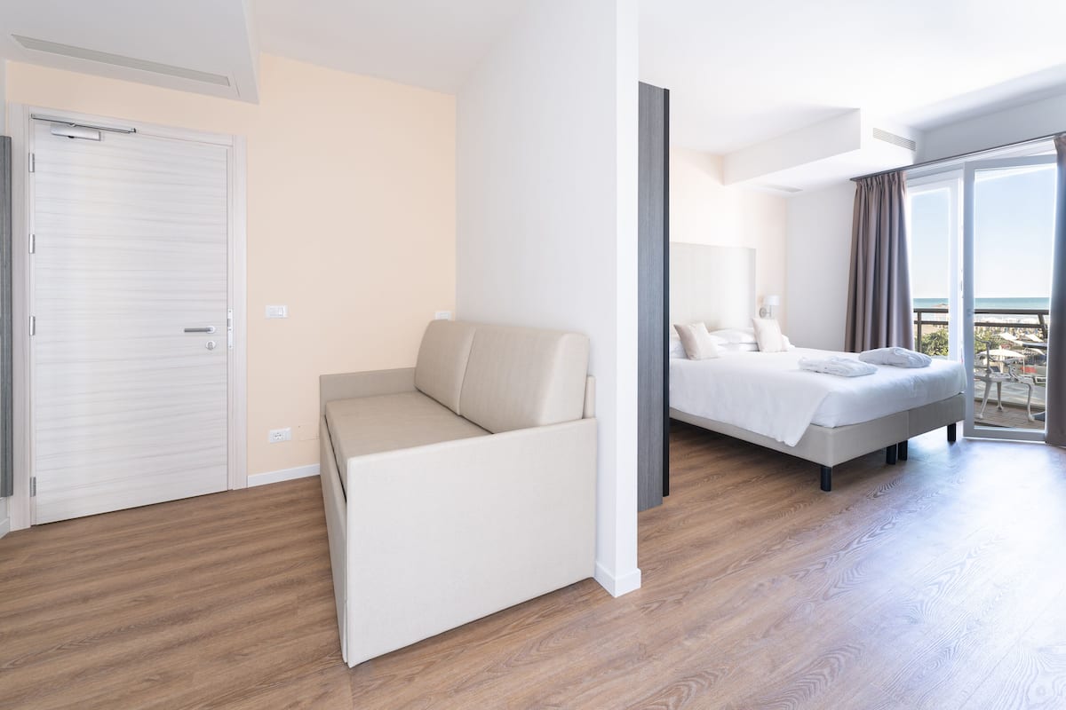A modern hotel room with a bed, a sofa, a sliding door, and a balcony with a view.