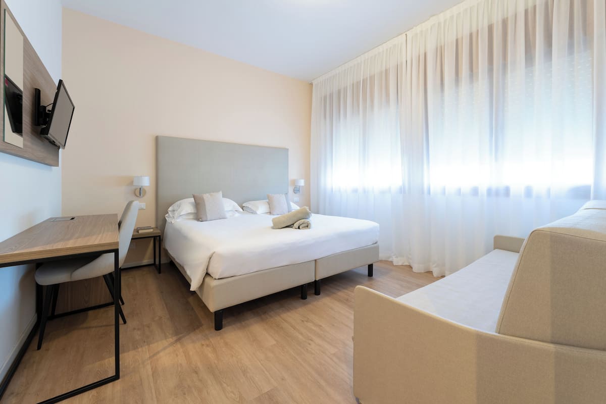 A hotel room with a bed, a desk, a chair, a TV, and a large window with curtains.