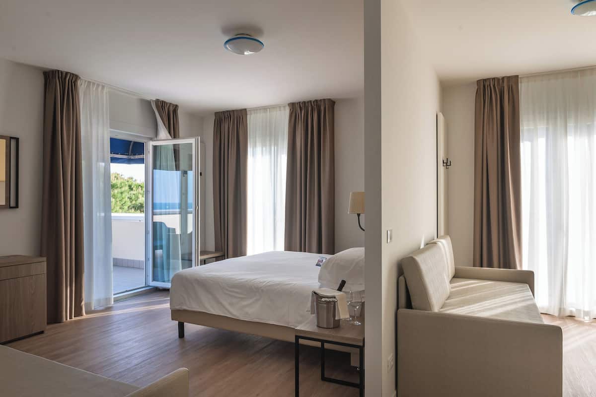 A hotel room with a bed, a sofa, a small table with a pitcher and glass, and a view of the outdoors through a sliding door.