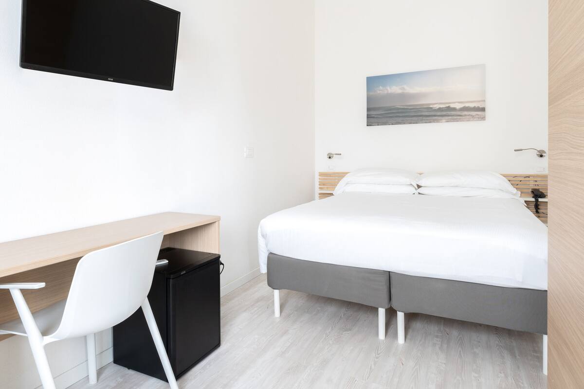 A modern hotel room with a bed, a desk, a chair, a television, and a painting on the wall.