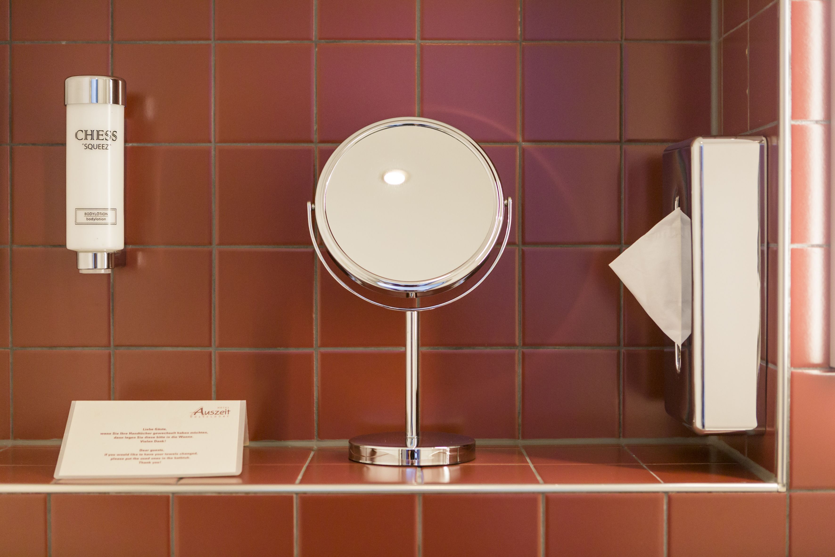 Un bagno con uno specchio rotondo, una bottiglia di lozione corpo CHIESS 'SQUEEZ', un dispenser di fazzoletti e un cartello con un messaggio.